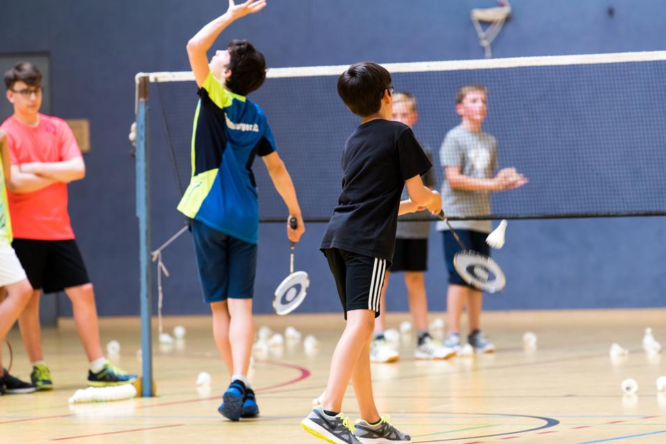 Gruppe von Kindern beim Badmintontraining in einer Sporthalle, mit Schlägern und Federbällen auf dem Boden.