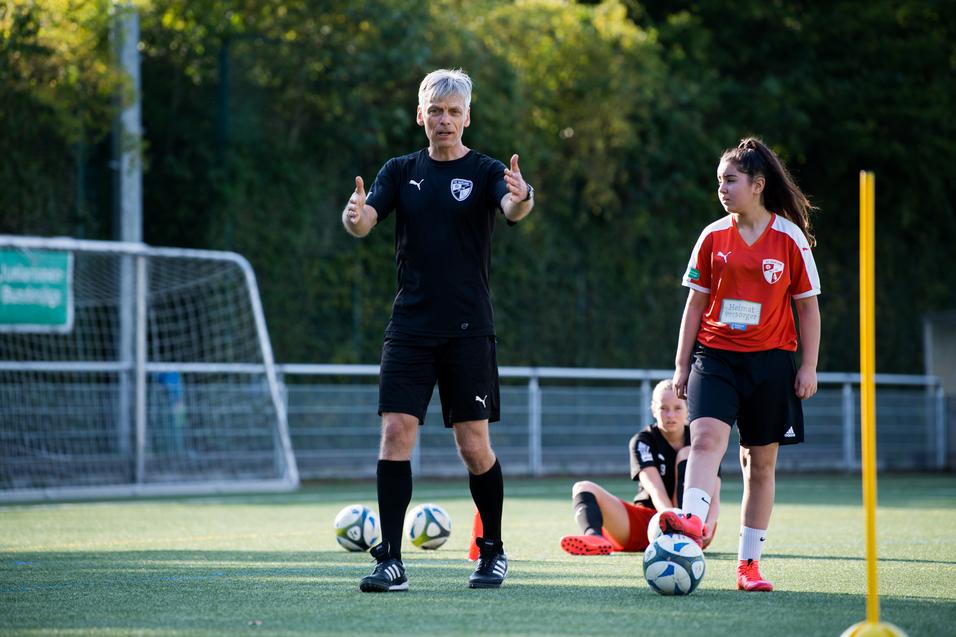 Trainer erklärt Technik beim Fußballtraining, während Spielerinnen auf dem Kunstrasen stehen und sitzen.