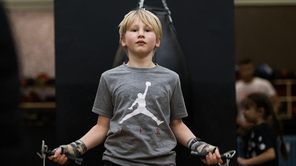 Junge mit blonden Haaren hält ein Springseil, im Hintergrund hängt ein Boxsack an einer Kette.