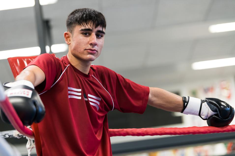 Jugendlicher Boxer in rotem Shirt lehnt an einem Boxring, trägt Handschuhe und schaut direkt in die Kamera.