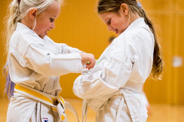 Zwei Kinder in weißen Karate-Anzügen helfen sich gegenseitig beim Binden ihrer Gürtel in einem Trainingsraum.