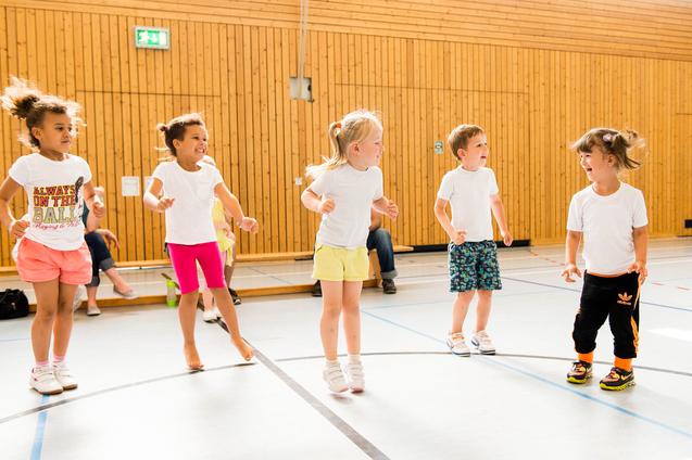 Fünf Kinder in T-Shirts springen fröhlich in einer Sporthalle mit Holzwänden und einen Zuschauer im Hintergrund.