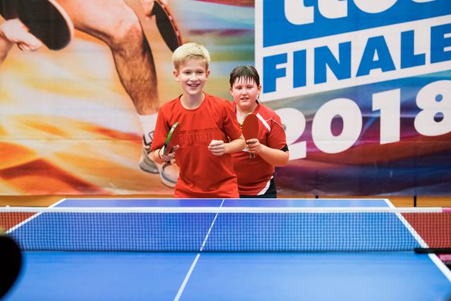 Zwei Kinder in roten Trikots stehen lächelnd an einem Tischtennistisch mit Schlägern in den Händen.