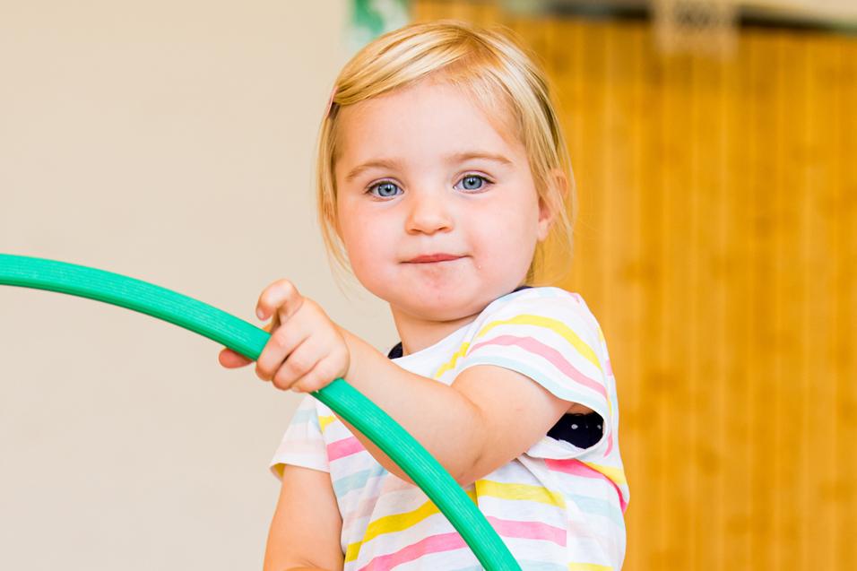 Kleines Mädchen mit blonden Haaren hält einen grünen Hula-Hoop-Reifen in einer Sporthalle mit bunten Reifen im Hintergrund.